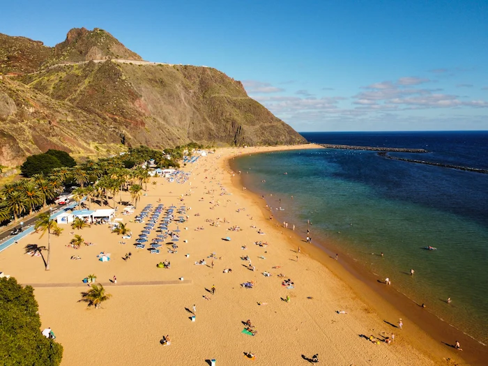 Aerial view of Playa de Las Teresitas with golden sand, umbrellas, and cliffs in the background.