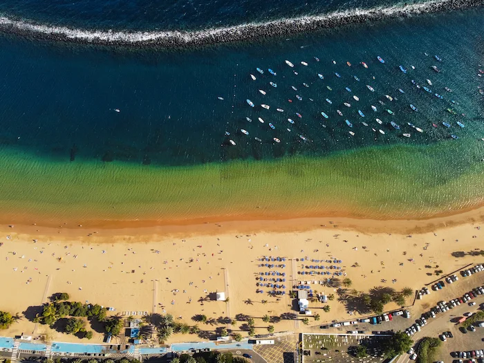 Top-down aerial view of Playa de Las Teresitas with breakwater and boats in the ocean.